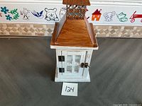 Front view of the candle holder showing the white distressed base, windowed door with metal latch, and wooden pyramid top with decorative metal accents