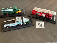 Three small Hess toy vehicles displayed on black plastic stands on a flat surface, with a handwritten number card 93.