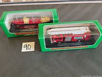 Two boxed Hess collectible mini vehicles positioned on a grey surface; left is the red Voyager ship from 2002, right is the red and white fire truck from 1999 with ladder.