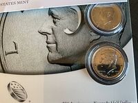 Photo showing two uncirculated Kennedy Half Dollar coins dated 2014 and 2014-D in clear protective capsules, on a background with a detailed close-up image of Kennedy's profile.