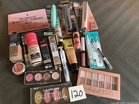 The photo displays a variety of unopened makeup products arranged on a flat surface with visible makeup brand names, types, and packaging distinct in shape, color, and design.