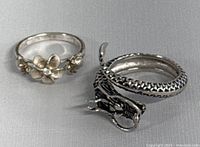 Two 925 silver rings shown together on a neutral background. Left ring with floral design and clear stone, right ring designed as a coiled dragon.