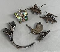 Photo showing all six pieces: two brooches (flower and bird), butterfly ring with abalone inlay, and silver dinosaur earrings