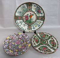 Three hand-painted Chinese ceramic pieces, including two plates and one platter, arranged with the platter on a stand behind the plates.