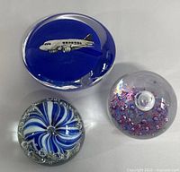 Top view of the 3 art glass paperweights showing airplane motif on blue glass, blue and white swirl with bubbles, and clear with purple/red floral design.