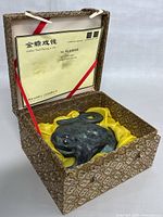Three-legged metal frog statue inside open decorative box with yellow cushion and certificate visible inside lid.