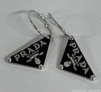 Front view of both silver hoop earrings with black triangular Prada logo charms laid flat