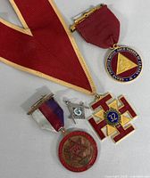 Several vintage Masonic medals and pins laid on a grey surface showing the full set with red ribbons and one tricolor ribbon