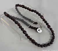 Garnet bead necklace shown fully extended over clear display stand, showing glossy dark red garnet beads and silver clasp.