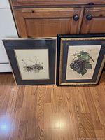 Two framed art prints leaning against a wooden cabinet on a hardwood floor, one showing an underwater scene with bunnies and the other a grape botanical print.