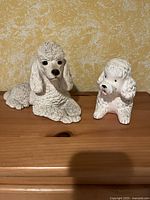 Front view image showing large white textured clay poodle figure with lying posture next to smaller smooth painted ceramic poodle figure