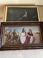 Two framed religious lithographs on a floor showing Jesus praying in a garden and Jesus in a wheat field with disciples.