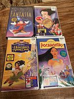 Four sealed Disney VHS tapes shown on wooden surface, titles Fantasia, Snow White and the Seven Dwarfs, The Hunchback of Notre Dame, Pocahontas