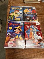 Four sealed Disney VHS tapes on wooden surface: Pinocchio Masterpiece Edition, Peter Pan Special Edition, Sleeping Beauty Special Edition, The Aristocats Gold Collection.