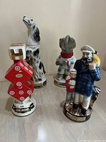 Four ceramic decanter figurines, including a sailor with child and dog, two different dog figures, and a red geometric dice stack.