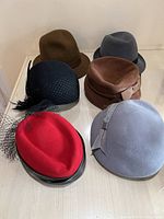 Top view of six vintage women's hats in various colors and styles: black with netting and feather, red with patent leather trim and netting, brown cloche style, navy blue, slate blue with bow, and grayish-blue.