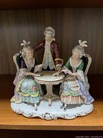 Full frontal view of the porcelain figurine group showing two women seated and one man standing around a white table with detailed decorations.
