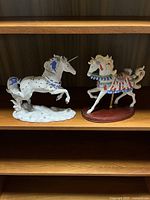 Shelf with two Lenox porcelain figurines: white unicorn with blue and silver details, and colorful carousel horse on wooden base.