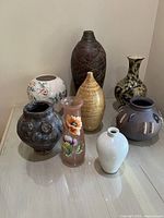 Photo showing the full group of 8 vases of varied materials, colors, and sizes arranged together on a table.