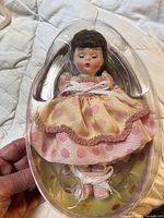 Full view of the doll inside original clear plastic egg-shaped packaging, showing the pink and yellow polka dot dress, white ribbon, and facial details.
