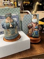 Two DeBrecht Studios music boxes, Father Frost playing trumpet and Merchant Santa with child, both on wooden bases.