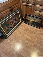 Two original oil paintings resting on a wood floor by cabinets, showing size and framing.