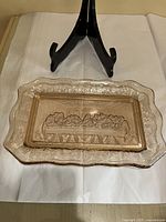 Pink Depression glass rectangular plate showing the entire piece with a black stand behind it on a white cloth, highlighting the scalloped grapevine border and Last Supper embossed scene in the center.