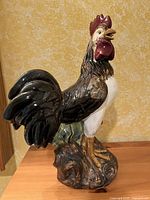 Side view of vintage ceramic rooster statue against a yellow patterned wall, showing full figure and base detail.