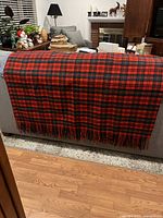 Red and black plaid Pendleton wool blanket draped over gray couch, showing full blanket and fringe detail.