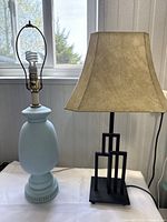 Photo showing the two lamps side by side on a table in front of a window. The blue ceramic lamp has no shade, the black geometric metal lamp has a tan fabric shade.
