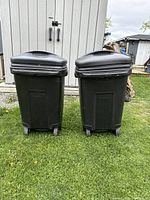 Two black plastic wheeled trash bins side by side on grass