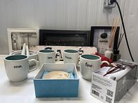 Photo showing four green inside white mugs labeled one through four, small jars, decorative shadow boxes, and brie warmer dish in a box.