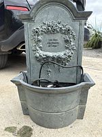 Front view of cast iron garden fountain showing decorative floral frame, inscription, and basin with water pump and power cord inside.