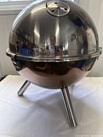 Front view of the shiny stainless steel dome lid charcoal barbecue grill supported by three tubular legs on a white surface.