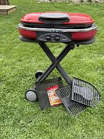 Red Coleman portable propane barbecue on black folding stand with wheels; cast iron grates, cooking basket, grill wipes displayed