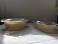 Two oval peach lustre glass casserole dishes with clear glass lids placed side by side on a white surface with light shadows.