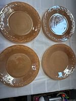 Four peach lustre FireKing plates arranged in a square, showing front surface including embossed laurel border.