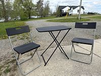 Two black folding chairs and one black folding table set outdoors on pavement and gravel background.