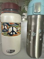 Photo of two drink containers side by side: a white GSI Outdoors water jug with colorful peace sign band and brown lid, next to a stainless steel Davids Tea travel mug with snowflake pattern and blue plastic lid.