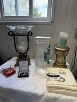 Photo showing Party Lite hurricane glass candle holder with metal base, large glass vase, gold-tone pillar candle holder with candle, leather wristlet, necklace in black box, and folded linen curtain.