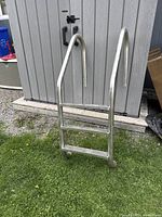 Steel inground pool ladder photographed from an angle, showing curved handrails and three steps with anti-slip surfaces.