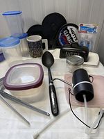 Photo showing entire group of assorted kitchen items including containers with lids, hand sanitizer bottles, spoon, tongs, butter dish, mug, and electric water dispenser.