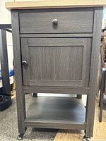 Front view of kitchen/bar cart showing the dark grey wood frame with a drawer and cabinet door, lighter wood tabletop, and bottom shelf with casters.