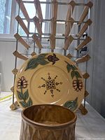 Photo shows three items: a wooden 24 bottle wine rack in the background, a cream-colored hand painted McNeil plate with floral green and purple designs in the middle, and a brown McCoy pottery dish #522 with textured pattern in the front.