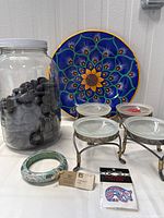 Photo showing the large jar full of dark river stones, blue and yellow sunflower wall plate, three pewter southwestern style tealight holders with glass bowls, native style bracelet bangle, and package of body art tattoos.