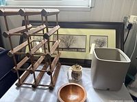 Photo showing wooden wine rack, framed tree picture, horsehair vase, wooden trinket bowl and white ceramic waste can near a window.