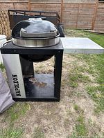 Full view of Napoleon charcoal kettle grill mounted in built-in stand with side shelf and Napoleon branding on stand.