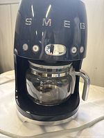 Front view of the black Smeg coffee maker with glass carafe seated on base. Shows the power, brew, and timer buttons and silver trim wear.