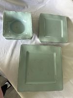 Photo showing square turquoise bowls stacked alongside square plates of two different sizes, laid on a white fabric.