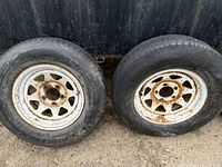 Two 205/75/R14 tires mounted on rusted white steel rims leaning against dark wall on dirt surface.